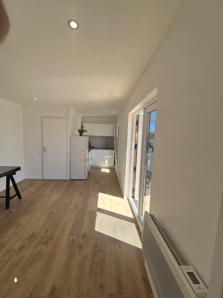 Séjour avec la cuisine au fond, sol aspect chêne et porte-fenêtre baignée de lumière