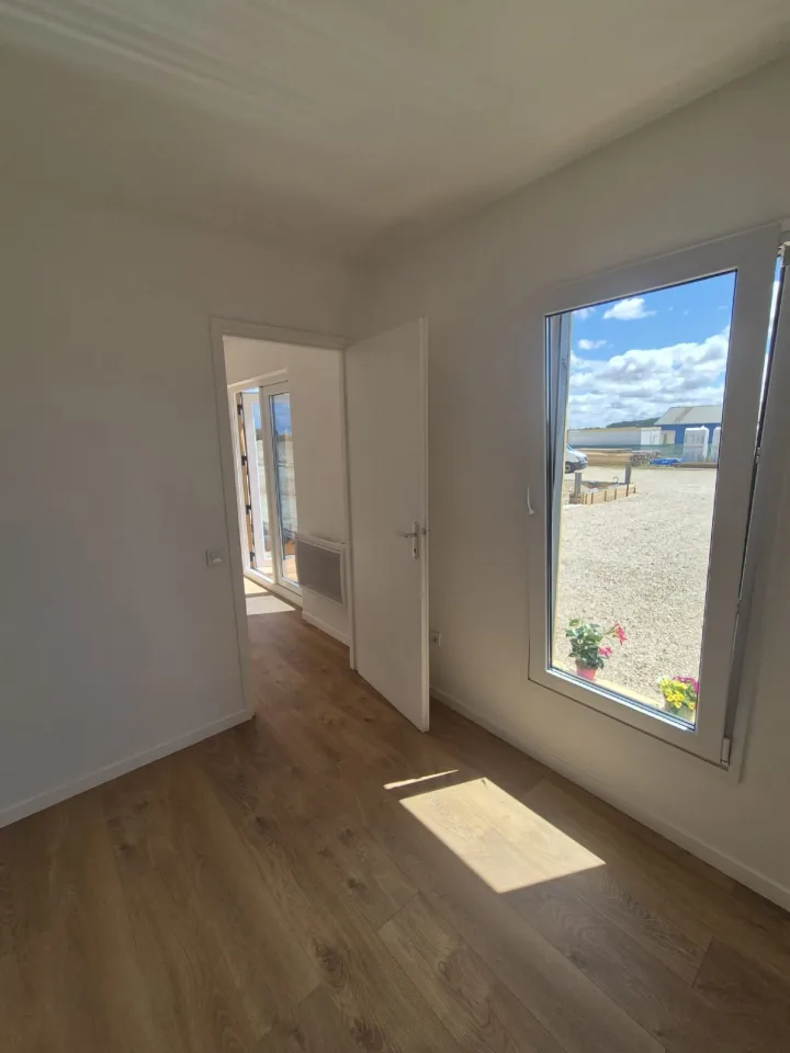 Pièce aux murs blancs et au sol aspect chêne, fenêtre sur la cour et porte ouverte vers le séjour
