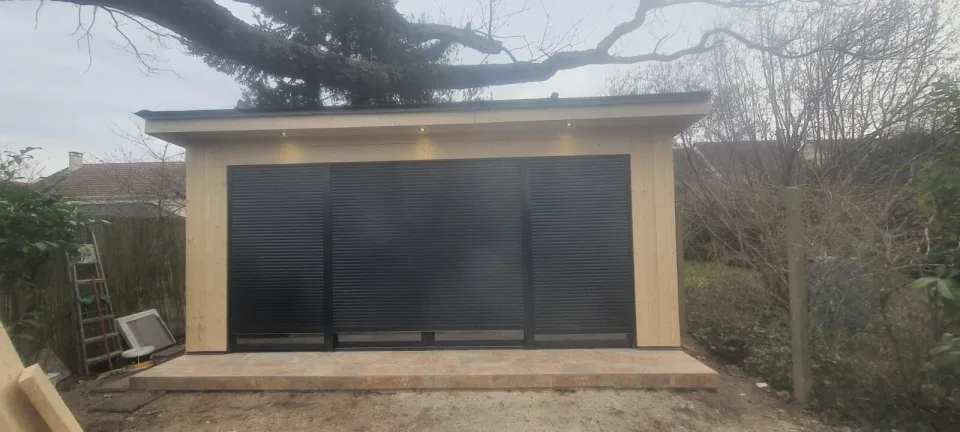 Volets roulants noirs fermés et spots encastrés allumés sous l’avancée de toit, abords du studio encore en travaux