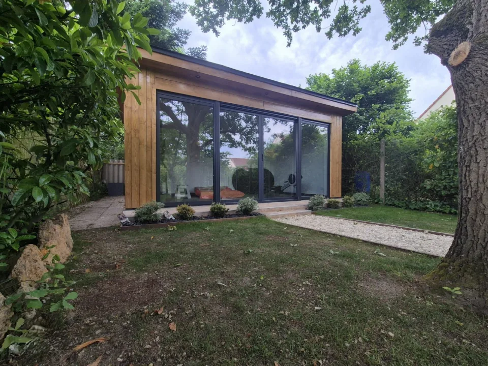 Studio livré, bardage vertical aux tons miel et grande baie coulissante vitrée à menuiseries anthracite, au milieu d’un jardin arboré