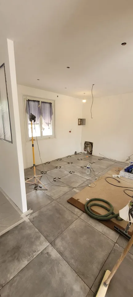 Pièce du rez-de-chaussée en travaux, grand carrelage gris en cours de pose avec ses cales de nivellement, projecteur de chantier sur trépied devant la fenêtre
