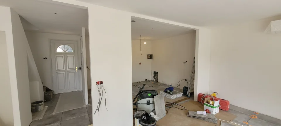 Entrée et porte d’entrée blanche à gauche, pièce en travaux au fond avec câbles en attente, aspirateur de chantier et cartons de carrelage