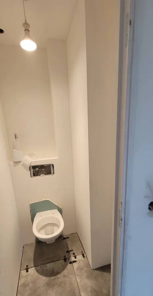 WC suspendu posé, bâti support encore visible par l’ouverture de la plaque de commande, carrelage de sol en cours de calage