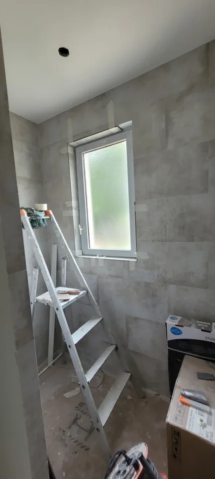 Salle d’eau aux murs couverts de grands carreaux gris, fenêtre à verre dépoli, escabeau et cartons de matériel au pied du mur