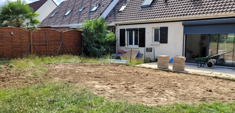 Façade arrière avant la terrasse, terre nivelée au premier plan, piles de dalles le long du mur et grande baie coulissante sous son coffre de volet roulant