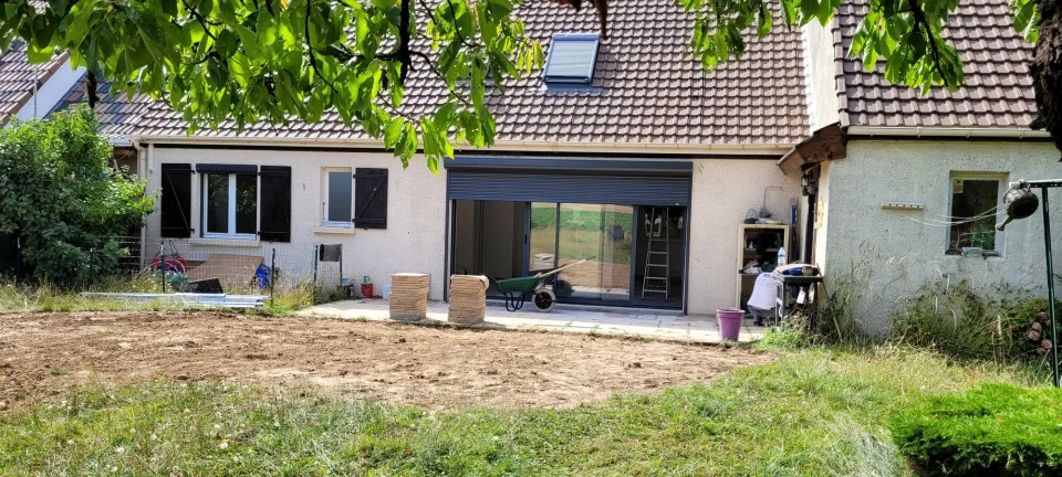 La maison vue du fond du jardin sous les branches, toiture percée de fenêtres de toit et sol mis à nu pour la future terrasse