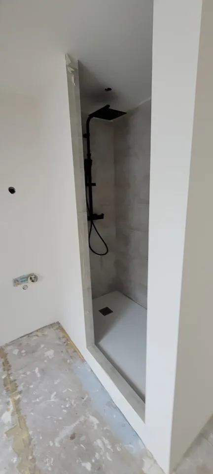 Douche avec colonne thermostatique noire et pomme de tête carrée, receveur blanc et murs carrelés, sol de la pièce encore brut