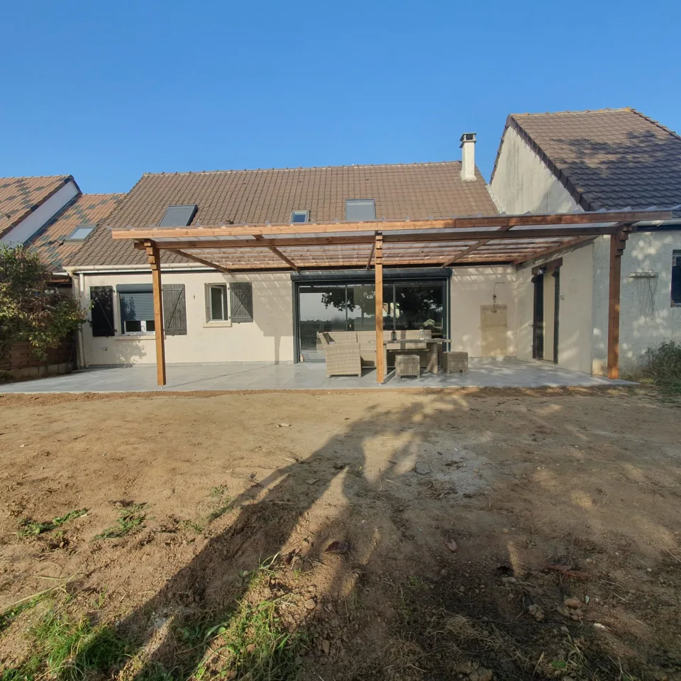 La façade arrière après les travaux, pergola en bois tout le long de la maison au-dessus de la nouvelle terrasse, terre nue au premier plan