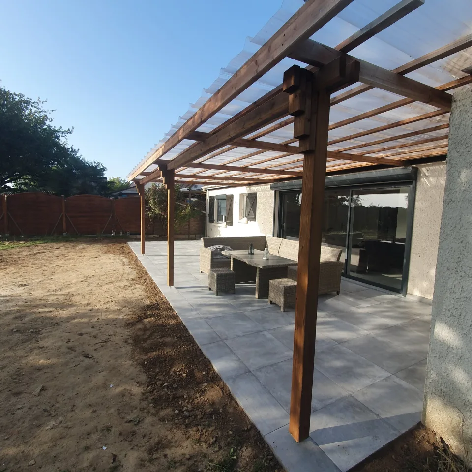 Sous la pergola, poteau et assemblage de poutres en bois au premier plan, couverture translucide et salon de jardin devant la baie coulissante