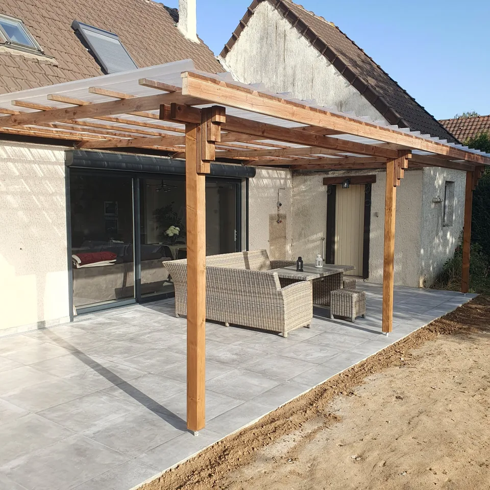 Poteau de la pergola au premier plan, salon de jardin tressé devant la baie coulissante et porte en bois au fond de la terrasse