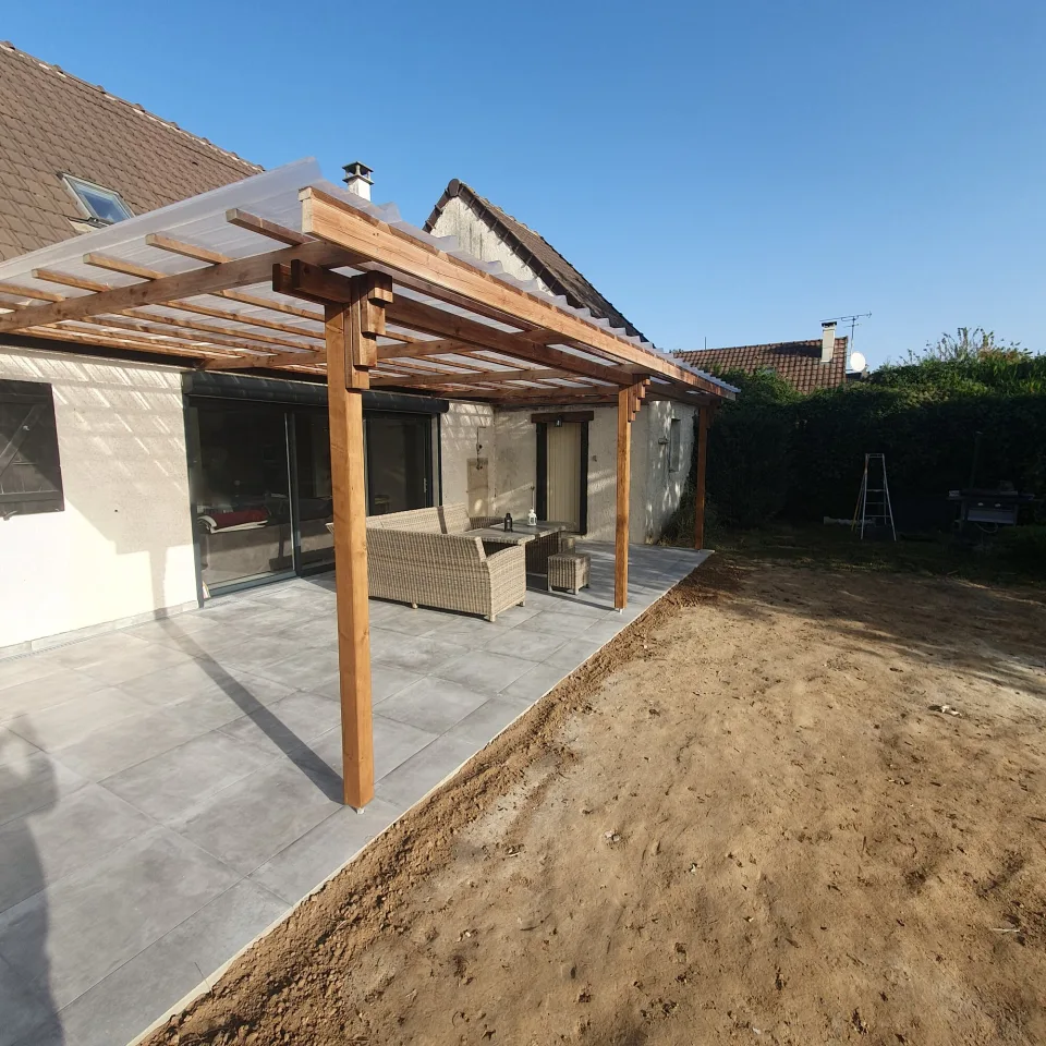 Pergola en bois et terrasse en grandes dalles grises vues en biais depuis le jardin, haie au fond sous un ciel bleu