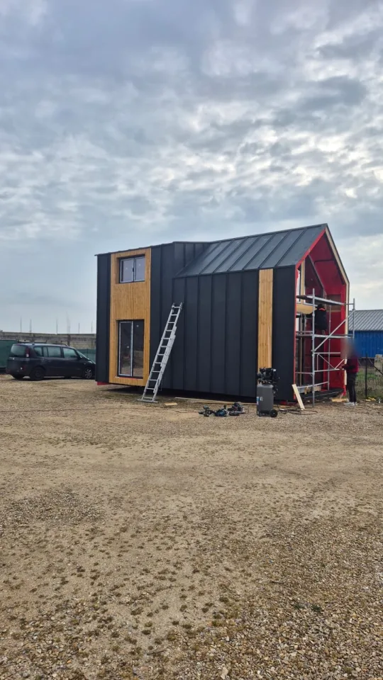 La maison sortie de l’atelier, bardage métal anthracite et lames de bois posés, pignon sous pare-pluie rouge derrière l’échafaudage