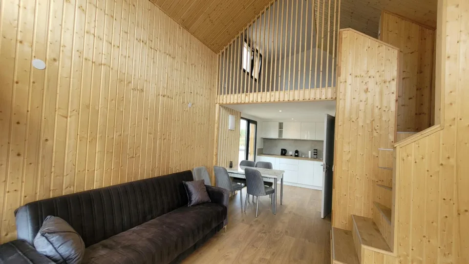 Séjour habillé de lambris clair, canapé, coin repas et cuisine sous la mezzanine au garde-corps à lames verticales