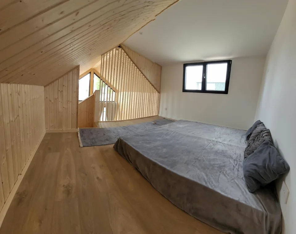 Chambre en mezzanine sous le rampant lambrissé, lit bas, fenêtre et garde-corps à lames ouvert sur le séjour