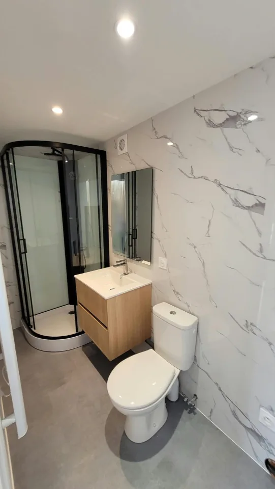 Salle d’eau, cabine de douche à profilés noirs, meuble vasque en bois, WC et murs effet marbre blanc