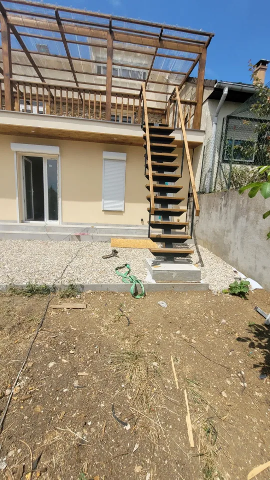 L’escalier extérieur à limon central en acier sur son socle en béton, bande de gravier au pied de l’extension et terre du jardin au premier plan