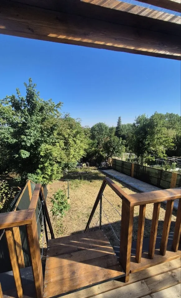 Depuis la terrasse, le haut de l’escalier et le garde-corps en bois, vue sur le jardin et les arbres