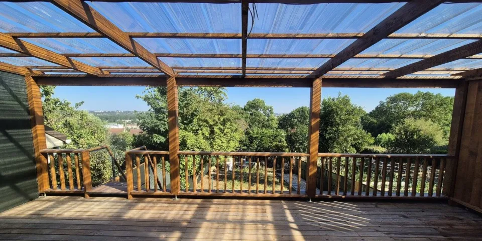 La terrasse sous la pergola à couverture translucide, platelage et garde-corps en bois, les arbres du jardin en fond