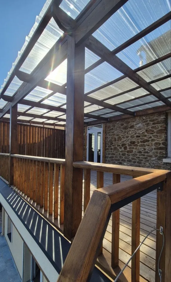 La pergola en bois contre le mur en pierre de la maison, garde-corps et porte-fenêtre donnant sur la terrasse