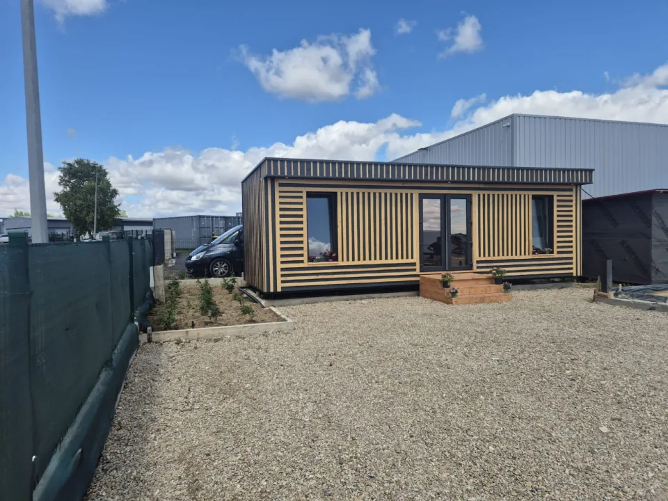 Tiny house livrée, bardage en tasseaux de bois naturel sur fond anthracite, porte-fenêtre, marches en bois fleuries et massif planté