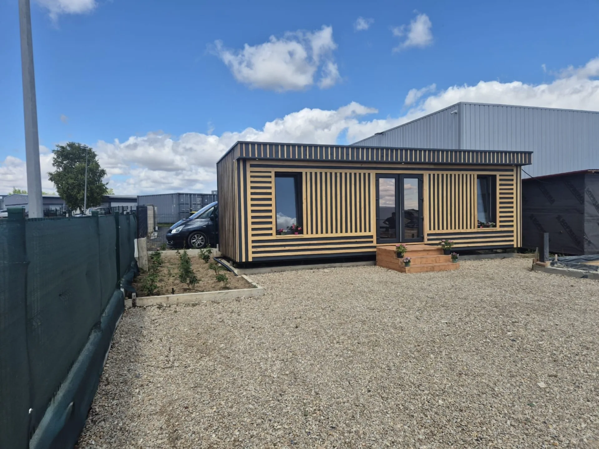 Tiny house livrée, bardage en tasseaux de bois naturel sur fond anthracite, porte-fenêtre, marches en bois fleuries et massif planté